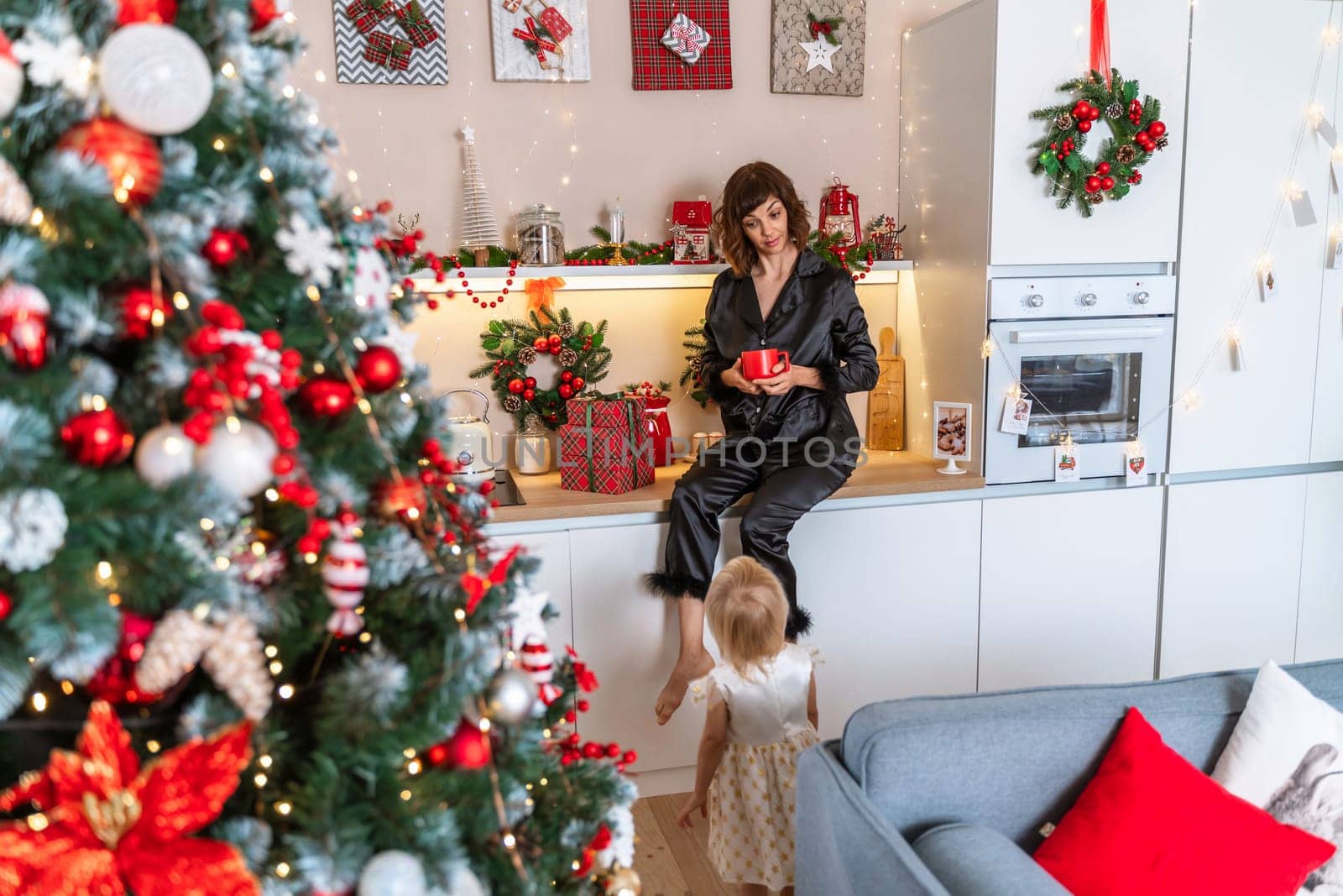 Christmas kitchen mom daughter. They are sitting on a table and drinks from a red mug in a decorated kitchen for christmas. by Matiunina