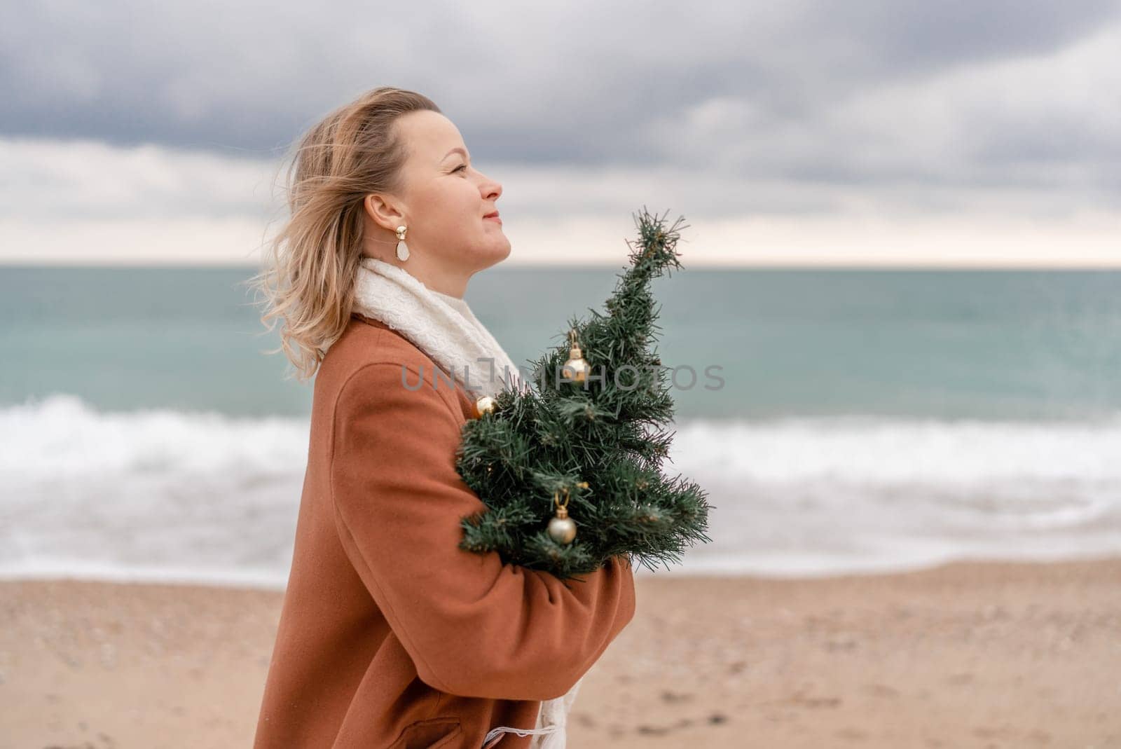 Blond woman Christmas tree sea. Christmas portrait of a happy woman walking along the beach and holding a Christmas tree on her shoulder. She is wearing a brown coat and a white suit. by Matiunina