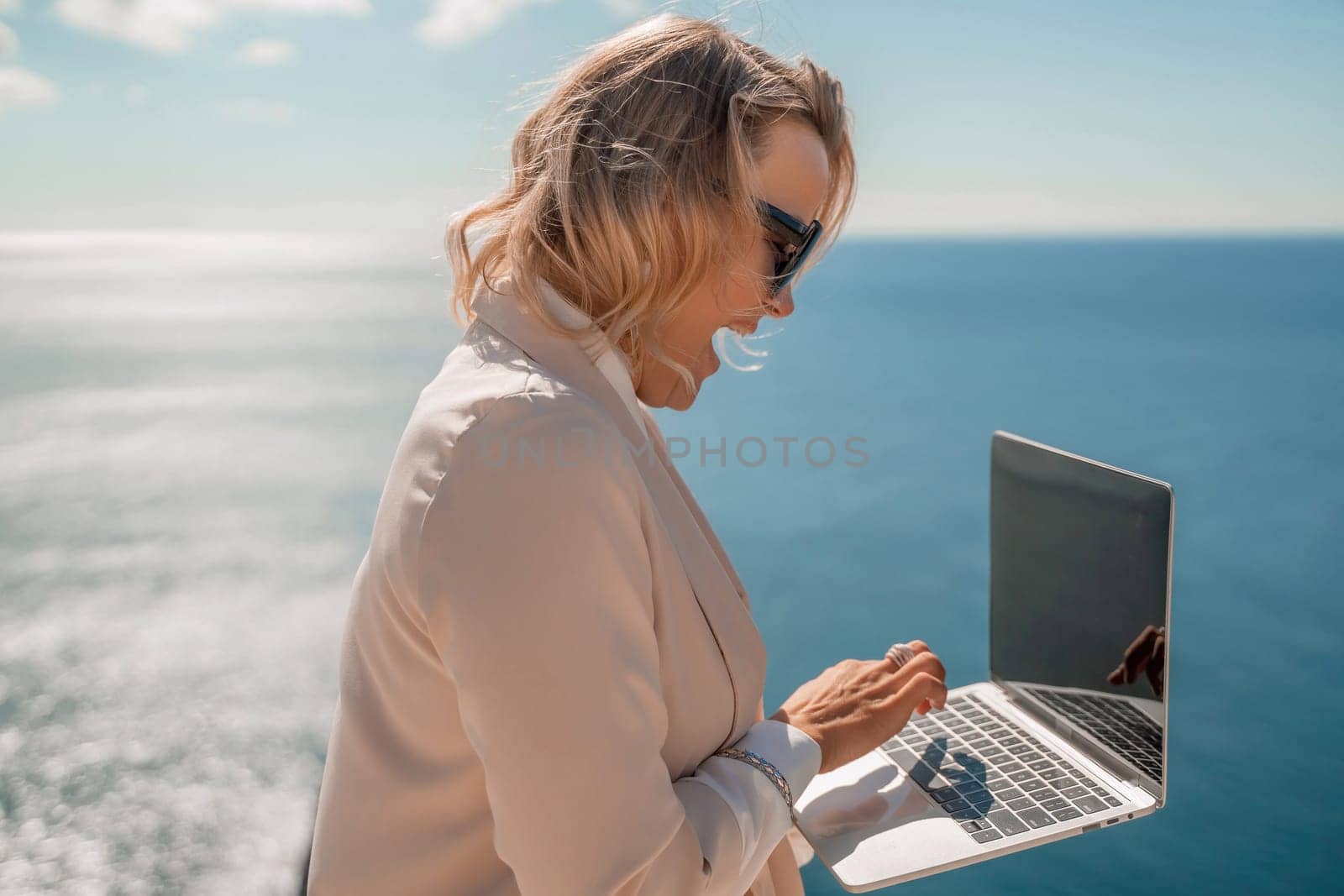 Freelance women sea working on the computer. Good looking middle aged woman typing on a laptop keyboard outdoors with a beautiful sea view. The concept of remote work. by Matiunina