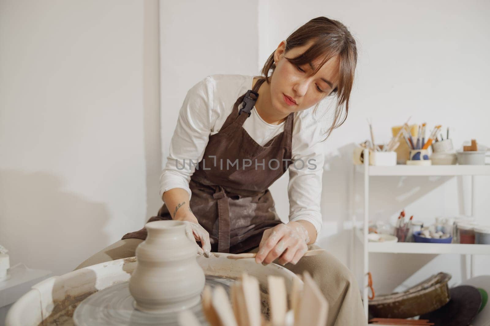 Young female artisan in apron sitting on bench with pottery wheel and making clay pot