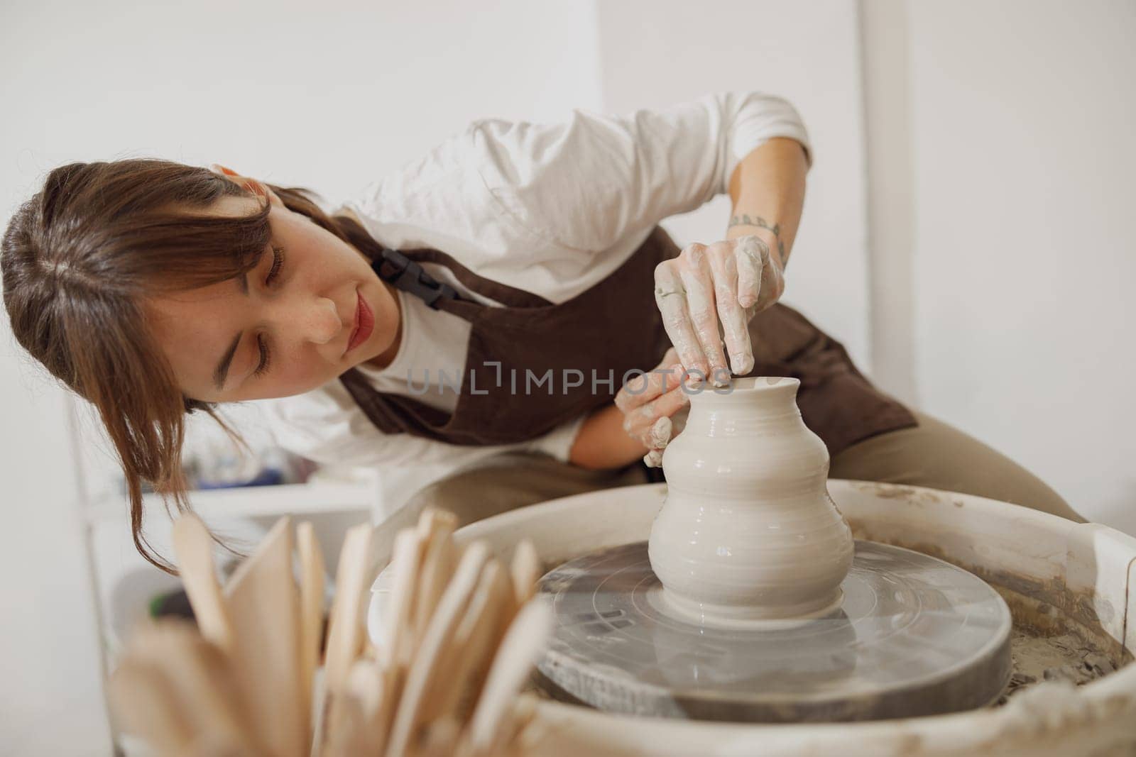 Smiling female artisan in apron sitting on bench with pottery wheel and making clay pot by Yaroslav_astakhov