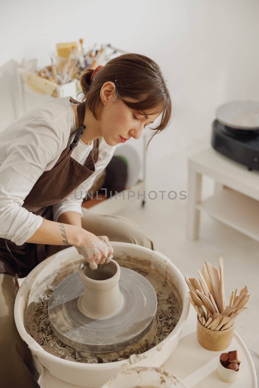 Smiling female artisan in apron sitting on bench with pottery wheel and making clay pot