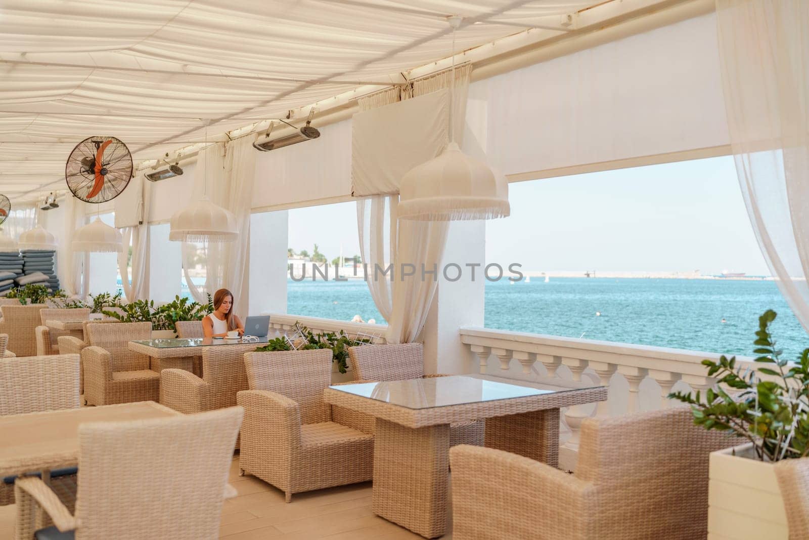 Woman coffee cafe laptop sea. Female freelancer using laptop on coffee break in modern cafe with sea view in the morning.