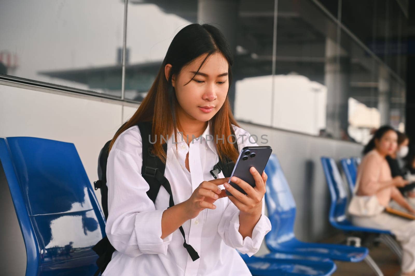 Young woman sitting at bus station, waiting for bus and using mobile phone to check bus schedules.