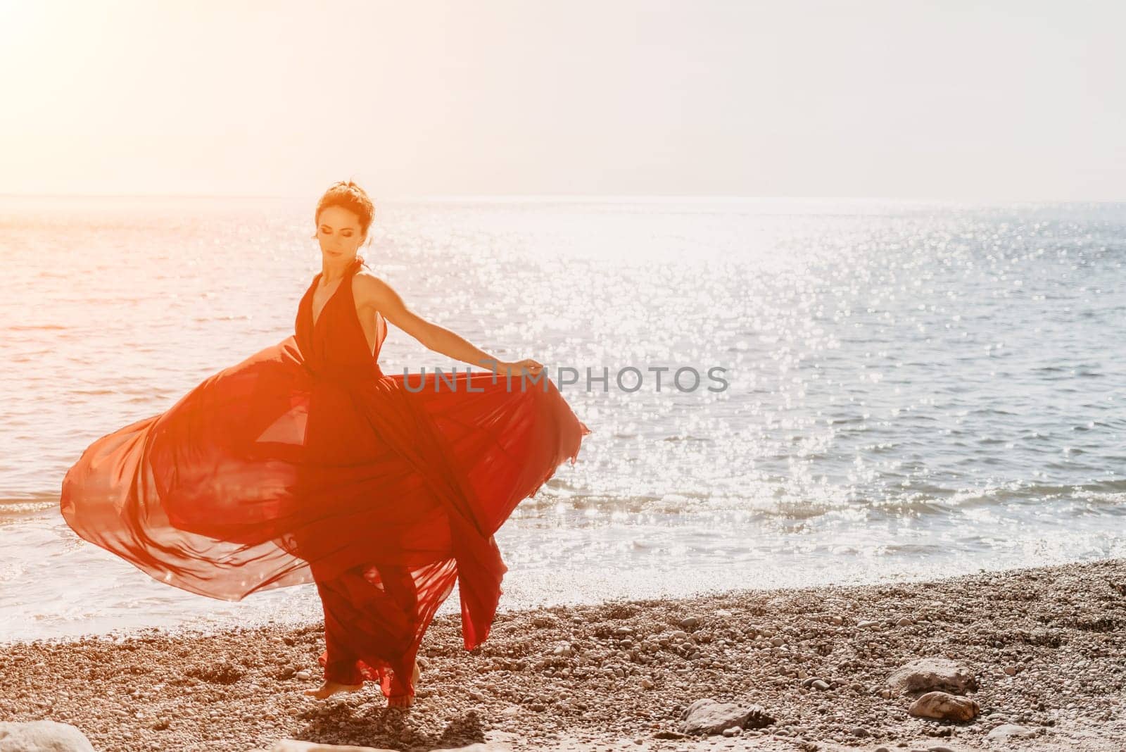 Side view a Young beautiful sensual woman in a red long dress posing on a rock high above the sea during sunrise. Girl on the nature on blue sky background. Fashion photo.