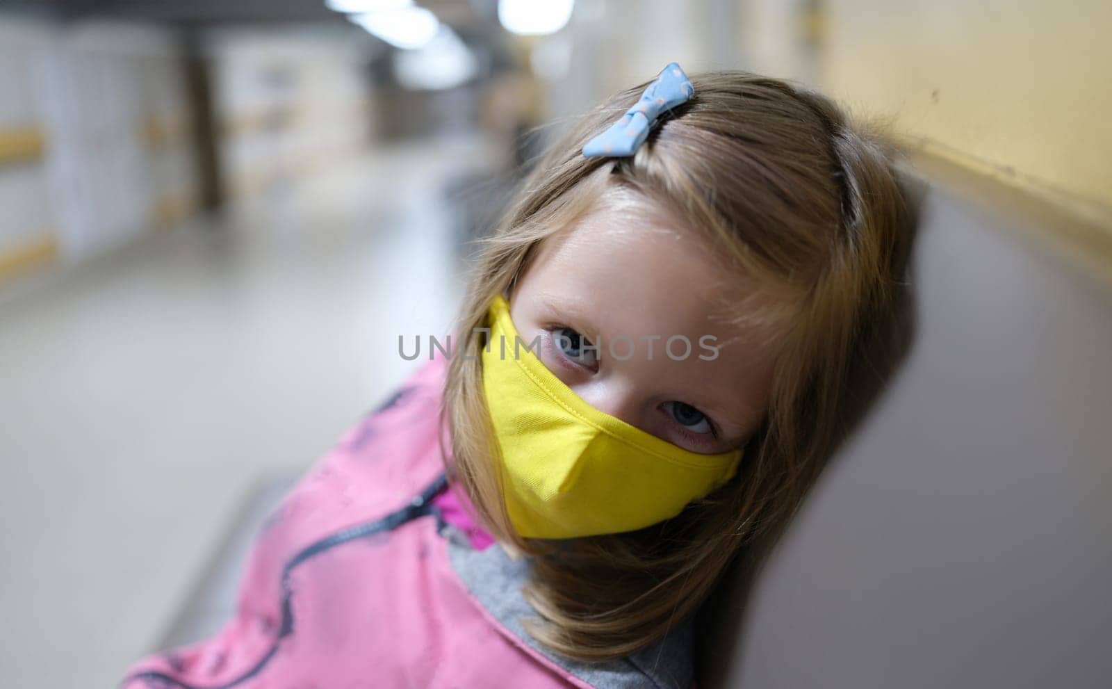Little girl in yellow protective medical mask sitting in hospital corridor. Medical care for children during covid19 pandemic concept