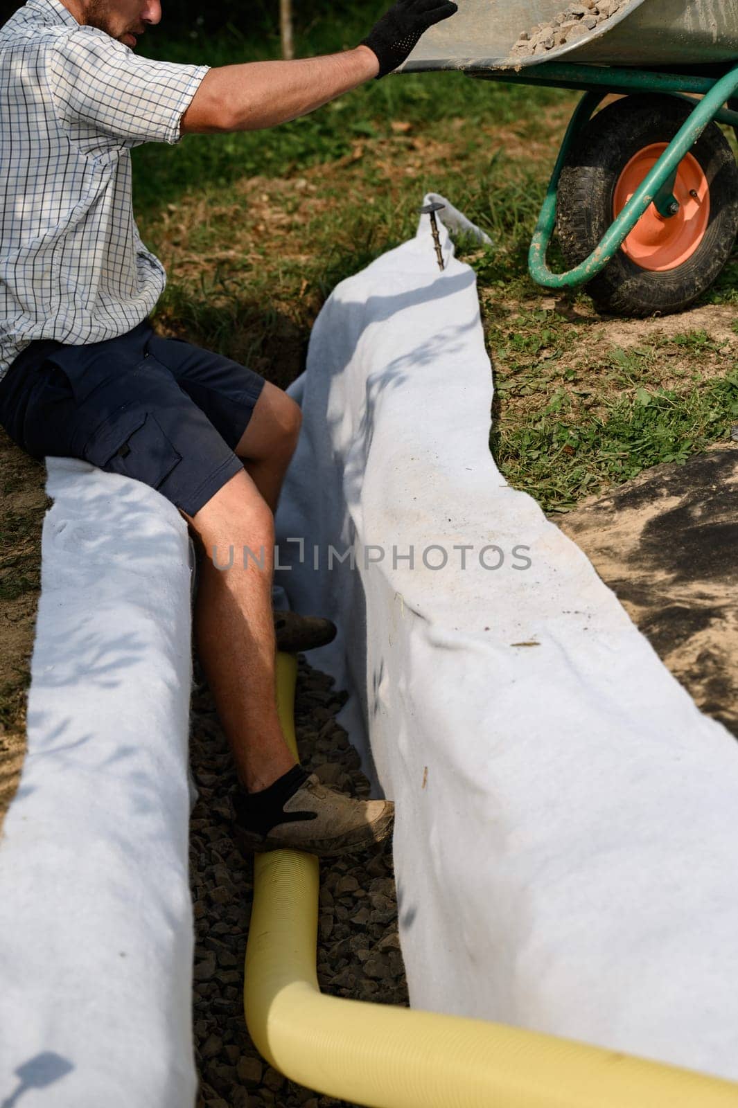 A worker stands in a drainage trench with white geotextile. Feet of the craftsman on the granite rubble closeup. by Niko_Cingaryuk