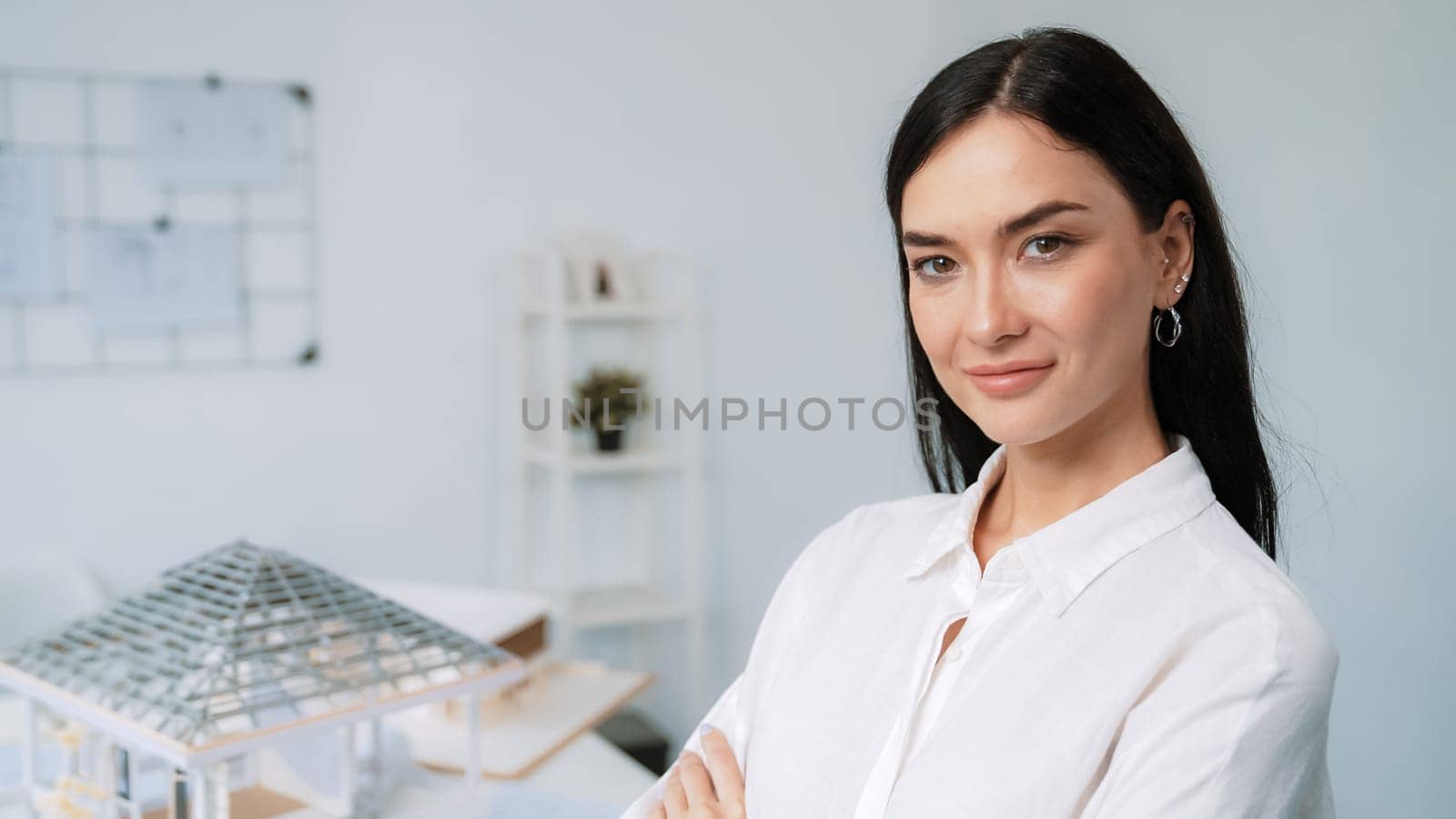 Close up portrait of young beautiful professional architect engineer cross arms and poses confidently with house model at white modern office with blueprint. Creative living and design. Immaculate.