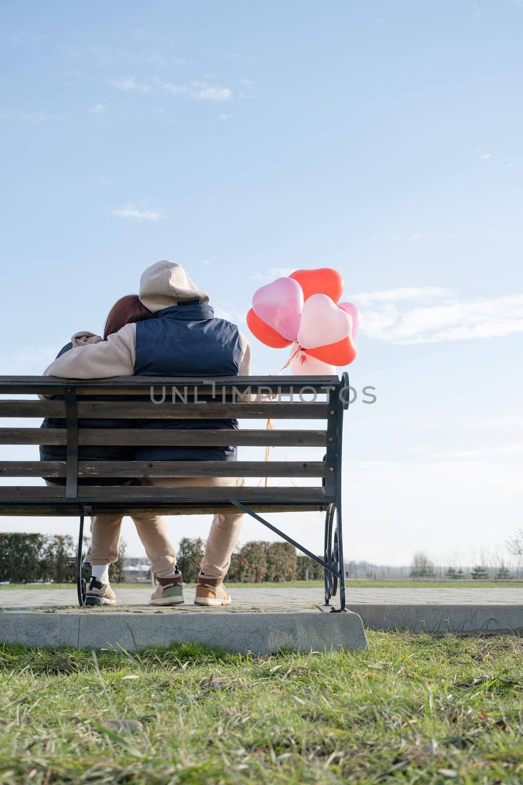 Outdoor photo of happy young woman enjoying date. cheerful romantic couple dating outdoors with balloons, sitting on bench, view from behind