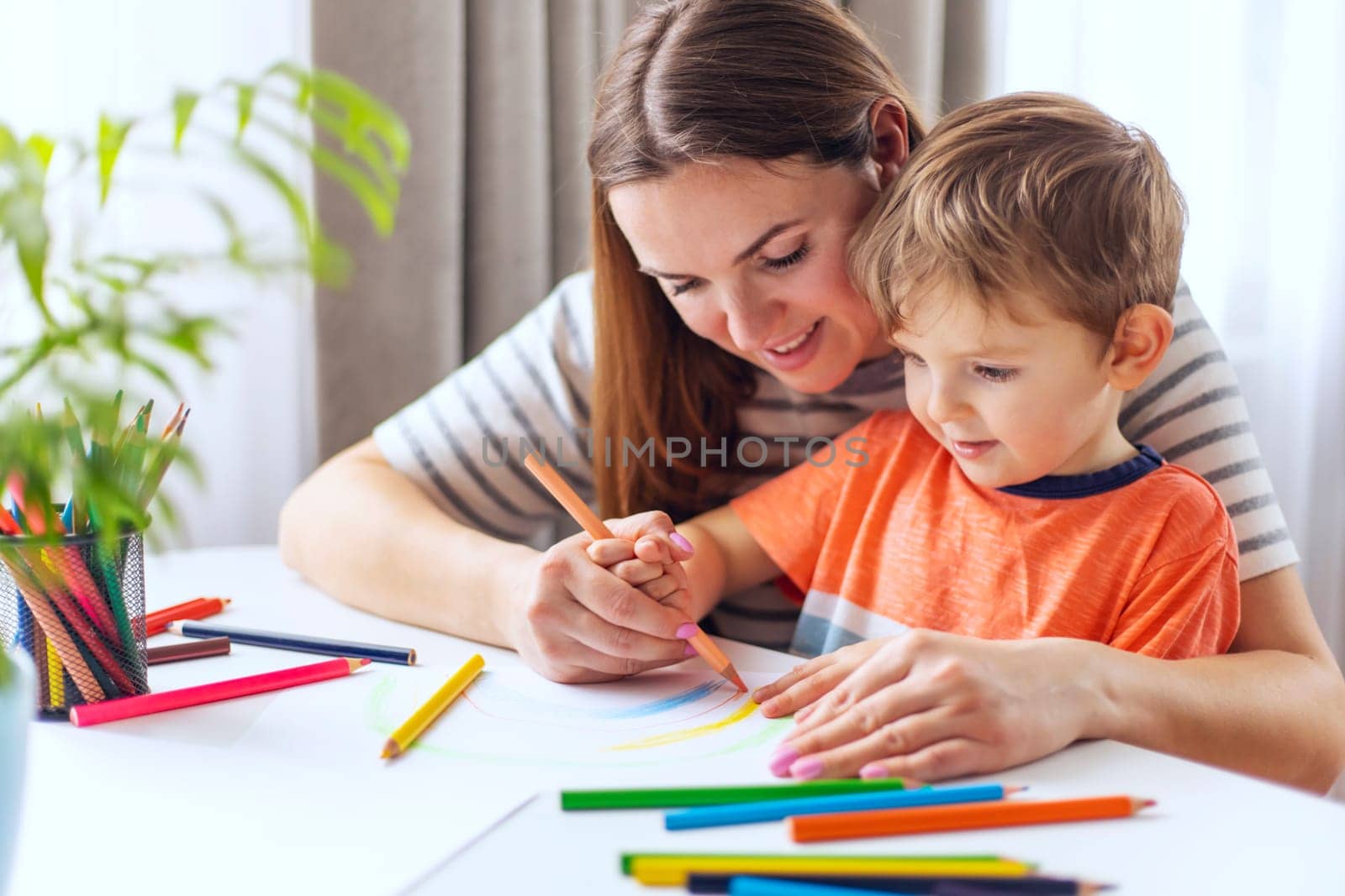 A caring mother guides her little boy in drawing with colorful pencils, creating a warm and educational home environment.