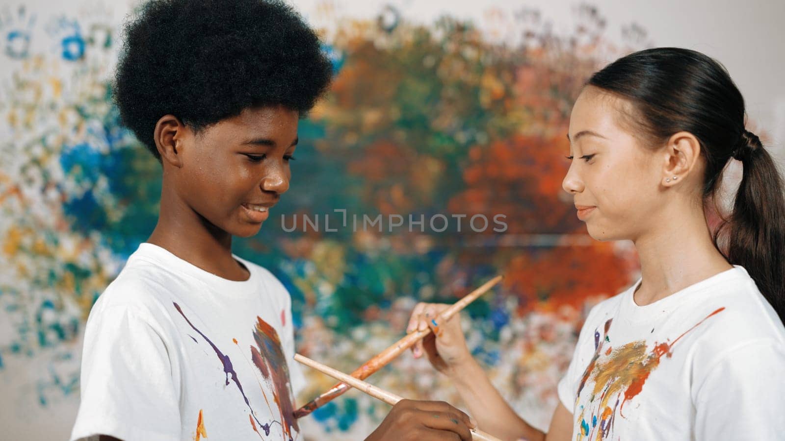 Smiling diverse children paint color on each other shirt at wall. Edification. by biancoblue