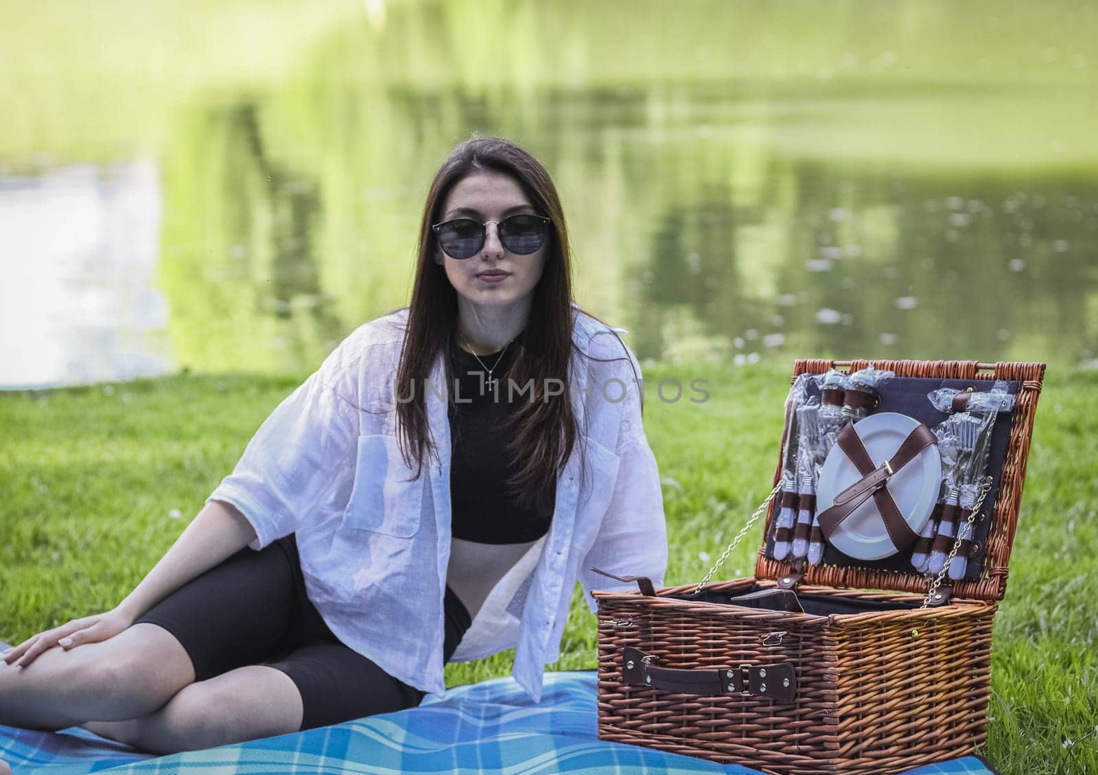 Portrait of a beautiful young caucasian brunette girl in sunglasses sitting on a plaid bedspread with a wicker basket relaxing in a park by the lake, close-up side view.PARKS concept and REC concept, picnic time, family vacation.