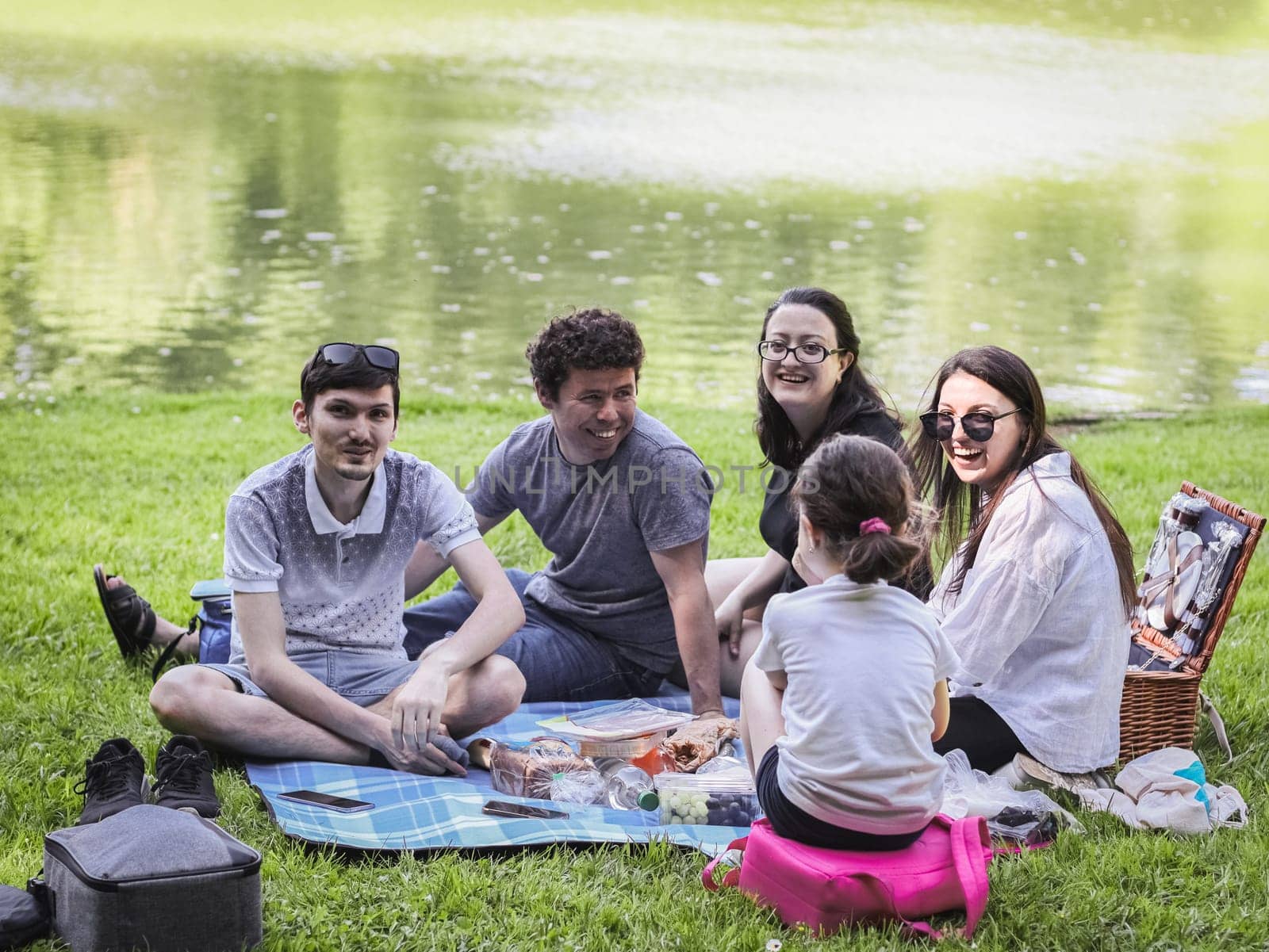 One young beautiful large Caucasian family with children sitting on a blanket with food, looking at the camera, laughing and having fun in a park near a lake on a clear sunny and spring day, close-up side view. Family picnic concept.
