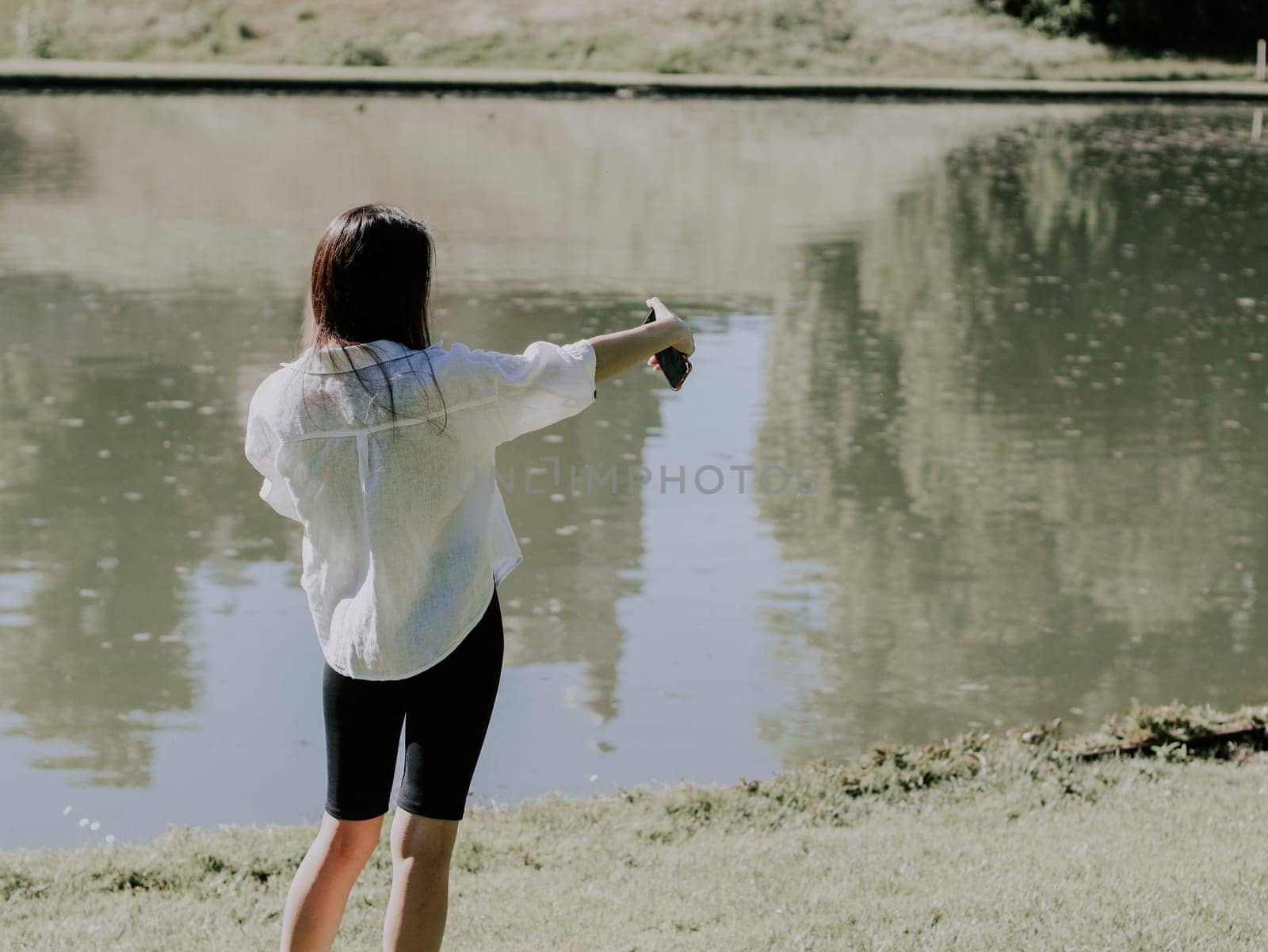 Portrait of a beautiful young caucasian brunette girl in a white shirt from the back makes a selfie on a smartphone while relaxing in the park by the lake, close-up side view.PARKS concept and REC concept, picnic time, using technology.