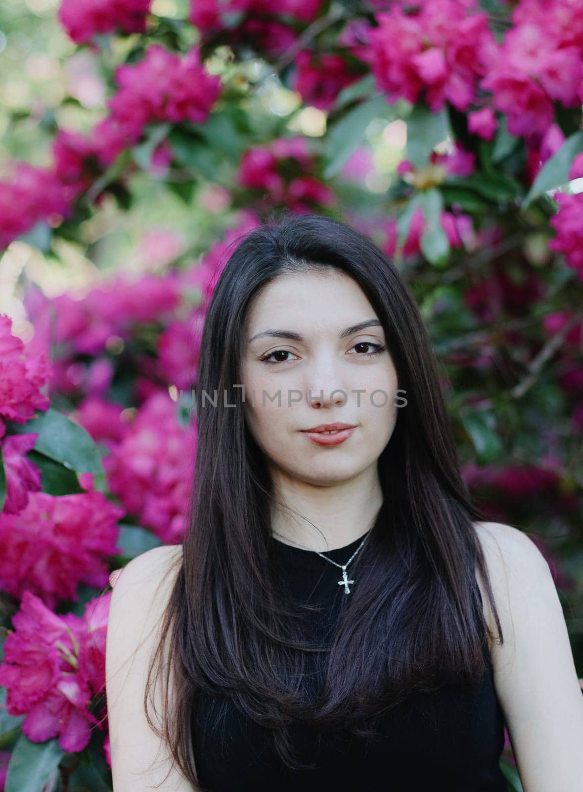 Portrait of one beautiful Caucasian brunette girl with flowing long hair in a black T-shirt stands near a flowering bush of pink rhododendron in a botanical park on a spring day, close-up side view with depth of field.