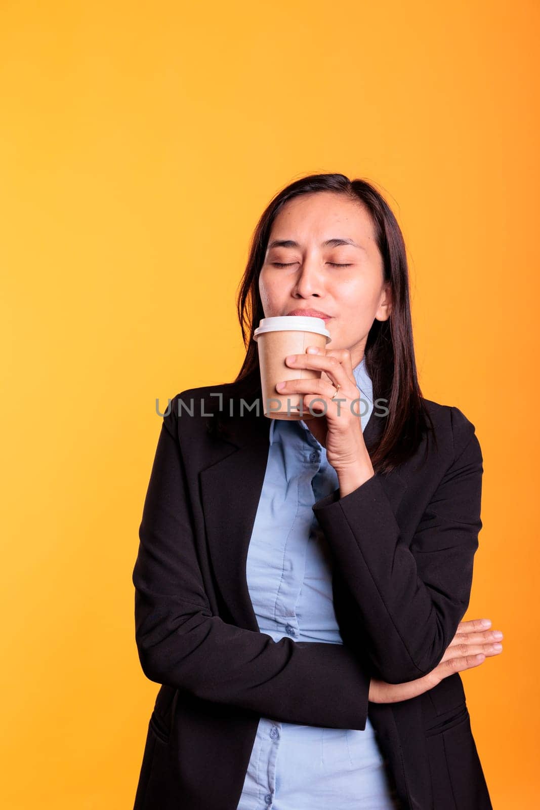 Smiling attractive woman enjoying drinking hot espresso in front of camera, posing in studio over yellow background. Smiling confident model in formal suit drinking hot beverage relaxing