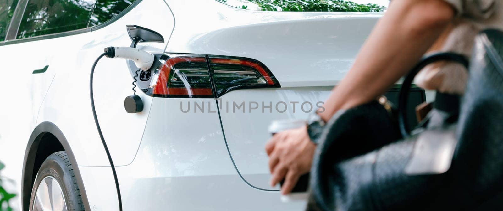 Young man recharge electric car's battery from charging station in outdoor green city park. Rechargeable EV car for sustainable environmental friendly urban travel. Panorama Expedient