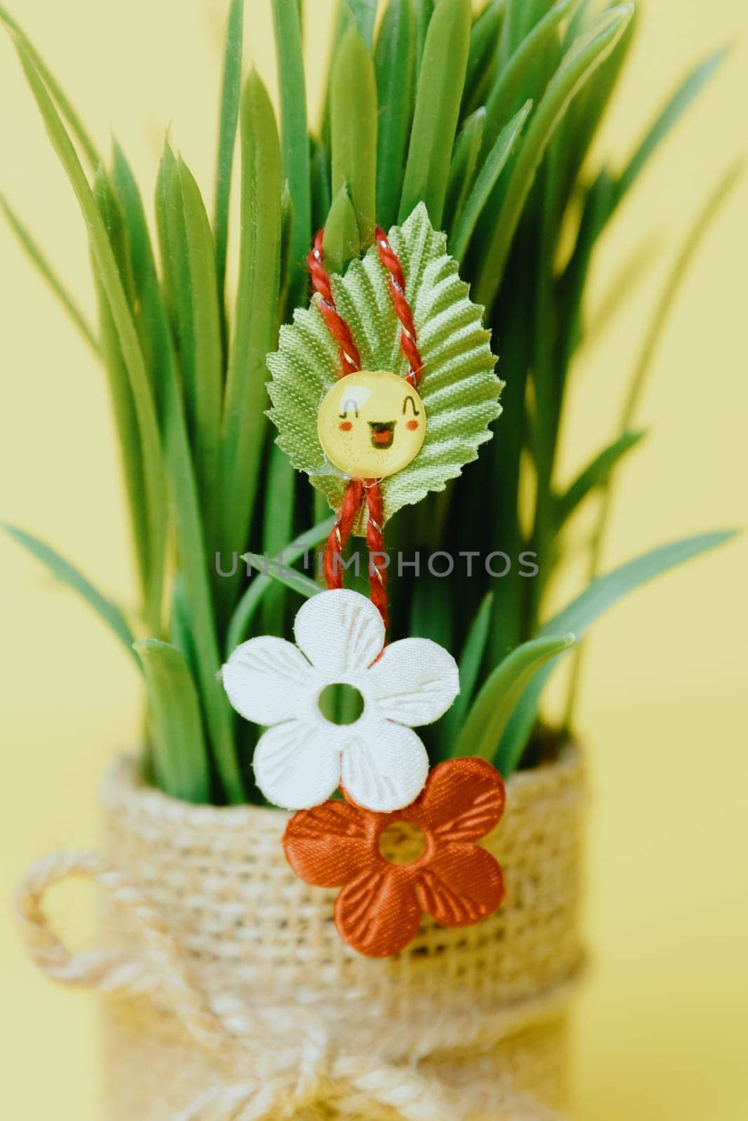 One beautiful homemade martisor of two flowers, a petal and a cheerful smiley face hangs on sprouted wheat in a jute pot on a pastel yellow background, close-up side view with depth of field.