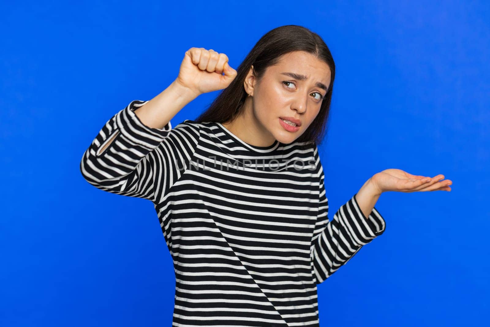Knock-knock who is there. Confused excited Caucasian woman knocking door gesture asking who is at home feeling embarrassed, no idea being clueless and uncertain. Young girl isolated on blue background