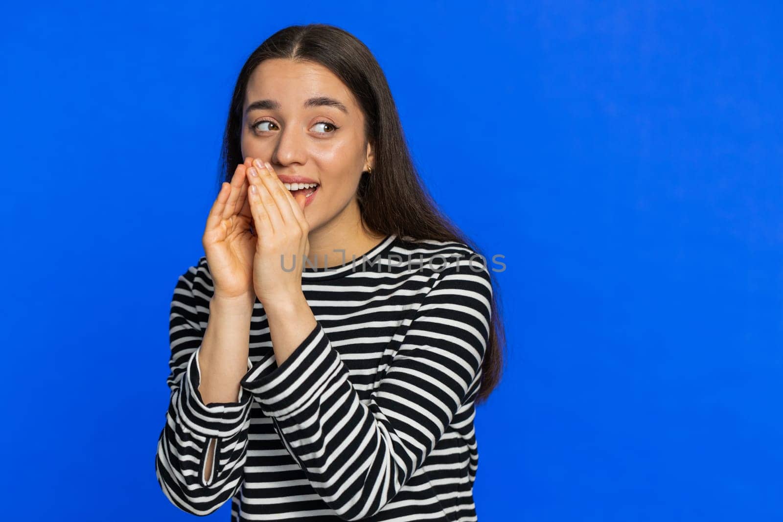 Keep my secrets, silence. Young woman whisper news rumors holding hands near mouth, share gossip, quiet. Girl telling interesting confidential story isolated on blue studio background. Copy-space
