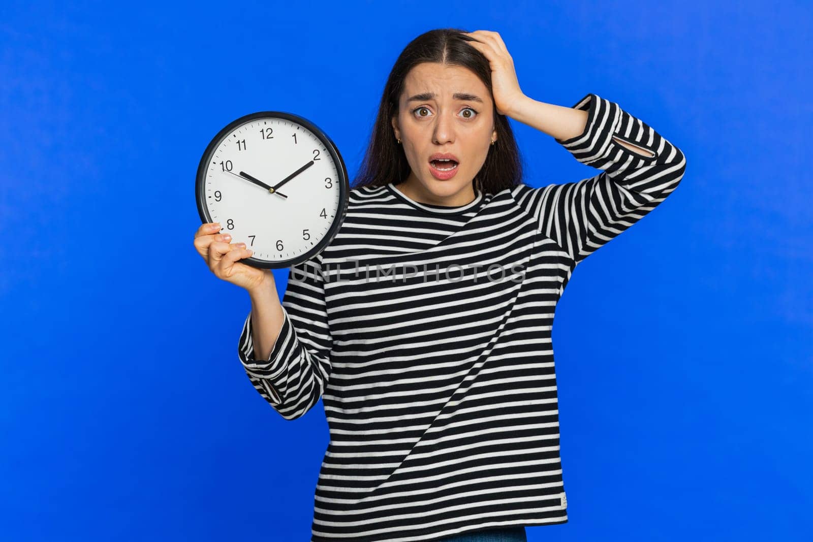 Caucasian woman with anxiety checking time on clock, running late to work, being in delay, deadline. Shocked girl looking at hour, minutes, worrying to be punctual isolated on studio blue background