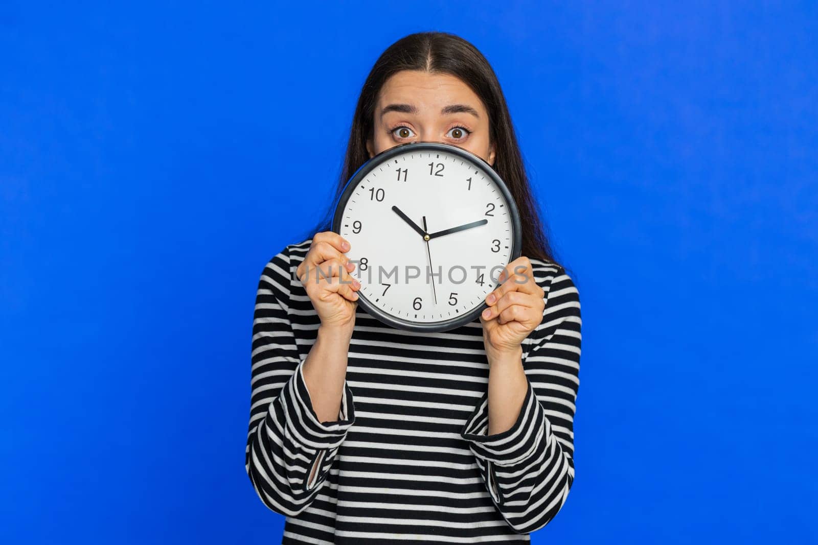 Serious strict Caucasian woman holding office clock hiding behind, checking time on watch, running late to work being in delay, deadline, obscuring face. Young girl isolated on blue background indoors