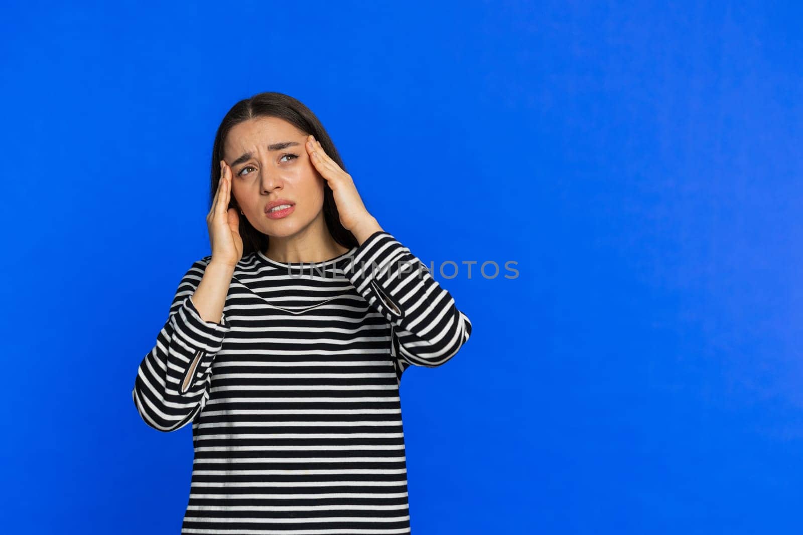 Exhausted young woman girl rubbing temples to cure headache problem, suffering from tension, migraine stress, grimacing in pain, high blood pressure isolated alone on blue wall background. Copy-space