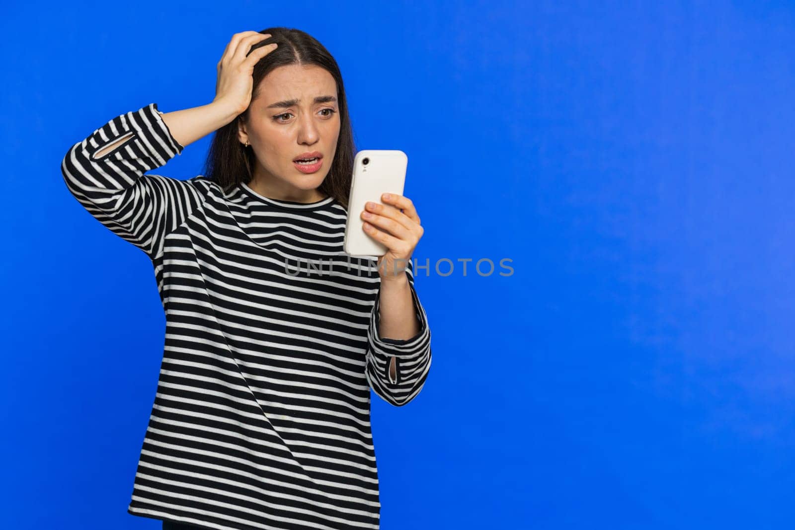 Upset sad Caucasian woman use smartphone typing browsing, play game, loses, surprised by sudden lottery results bad fortune loss fail unlucky news. Pretty girl isolated on blue background. Copy-space