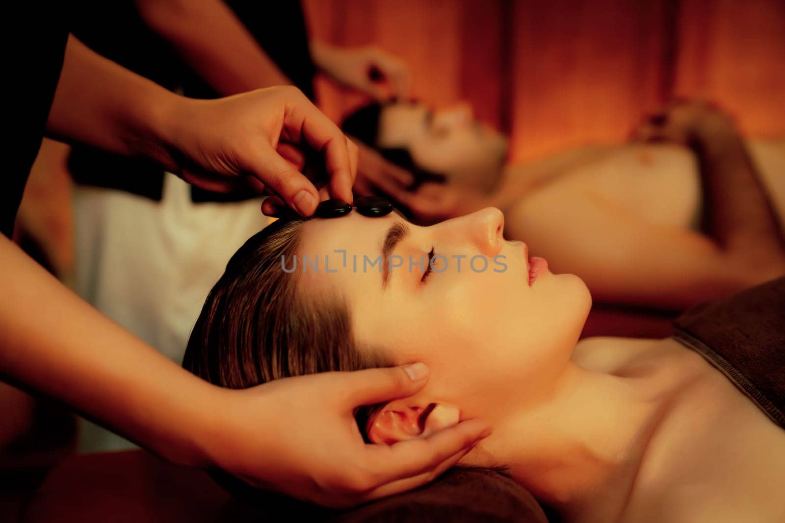 Caucasian couple enjoying relaxing anti-stress head massage with hot stone and pampering facial beauty skin recreation leisure in warm candle light ambient salon spa in resort or hotel. Quiescent