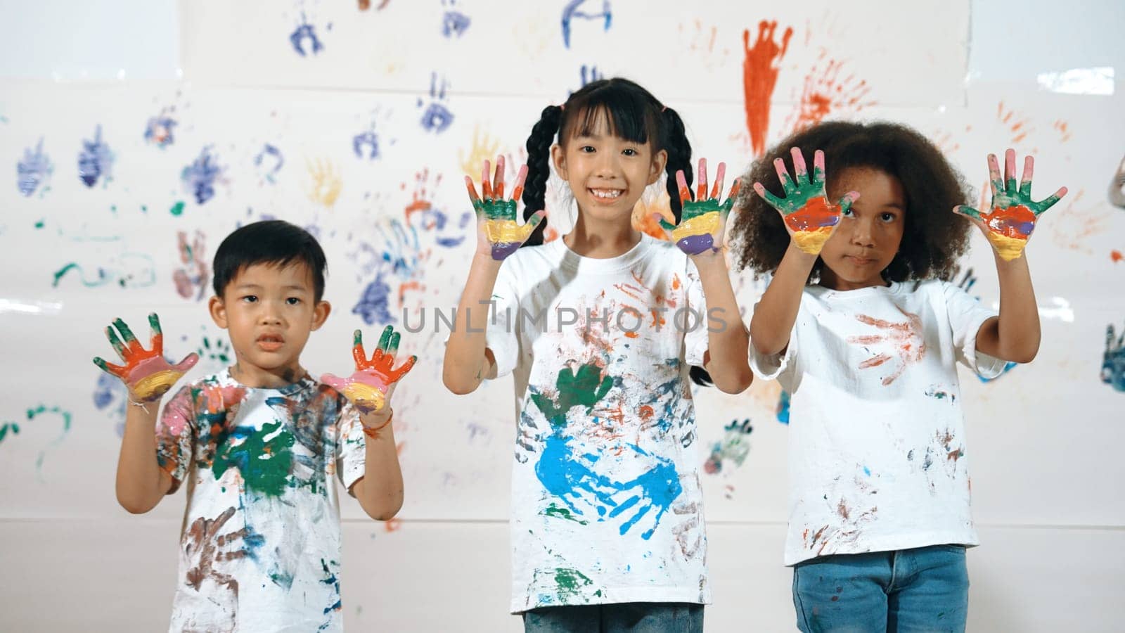 Diverse students put hands up together show colorful stained hands. Group of multicultural learner standing at white background with stained hands while looking at camera in lively mood. Erudition.