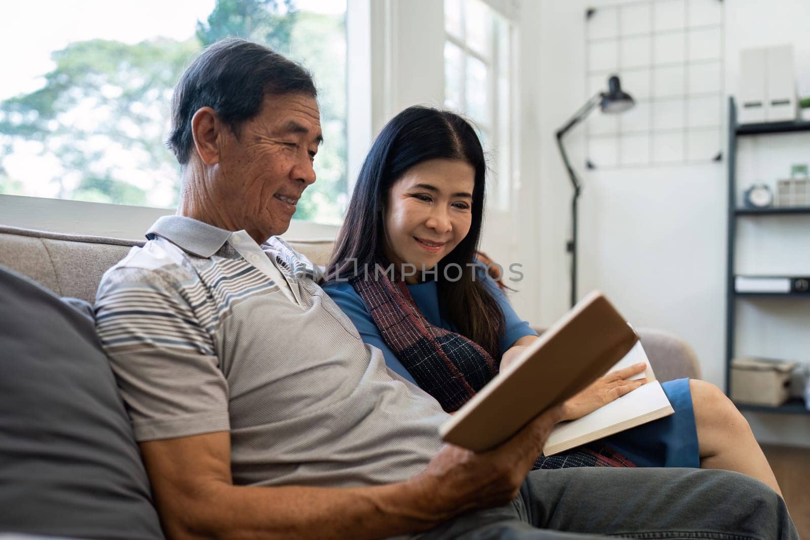 Retired elderly couple sits on couch in their home reading relaxing book. Senior Activity Concept.