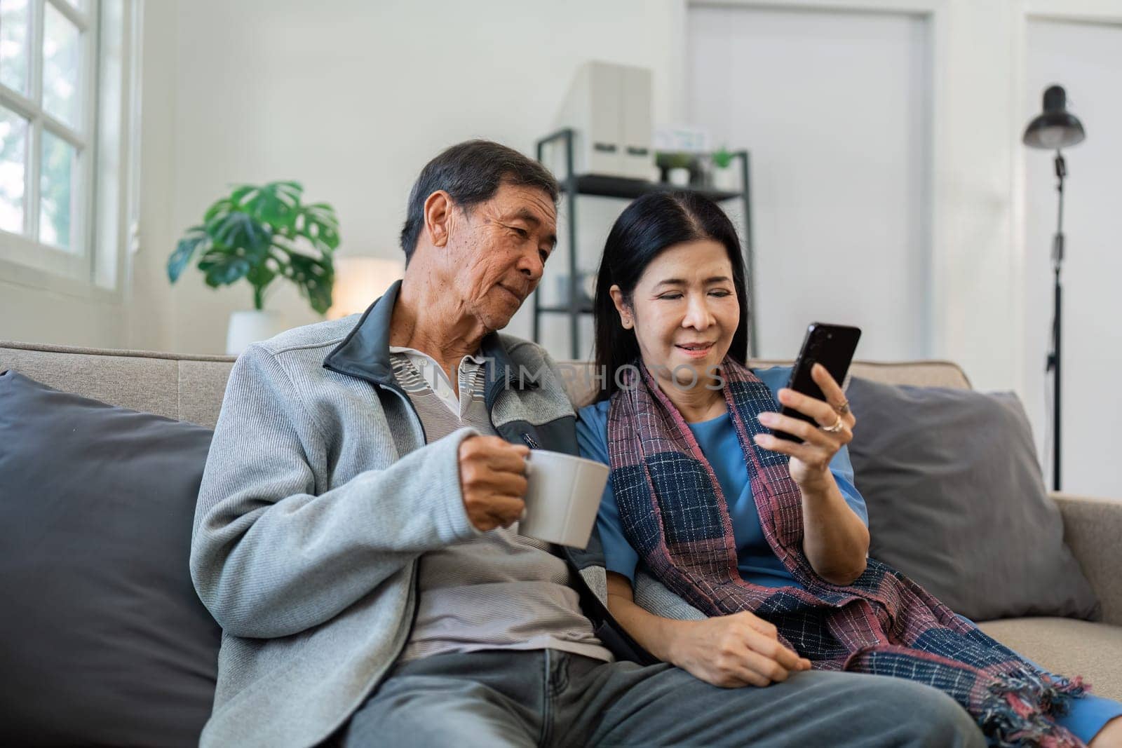 Retired elderly couple sits on couch drink tea and using mobile together and relax in their home. Senior Activity Concept by itchaznong
