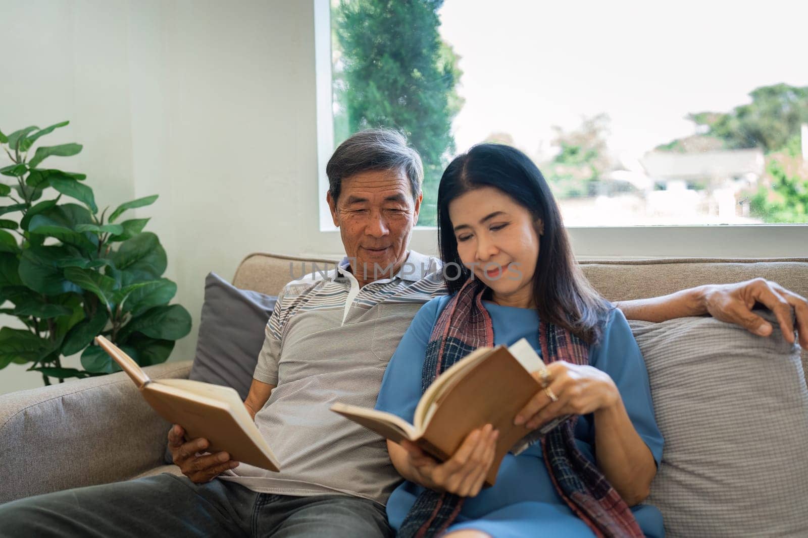 Retired elderly couple sits on couch in their home reading relaxing book. Senior Activity Concept by itchaznong