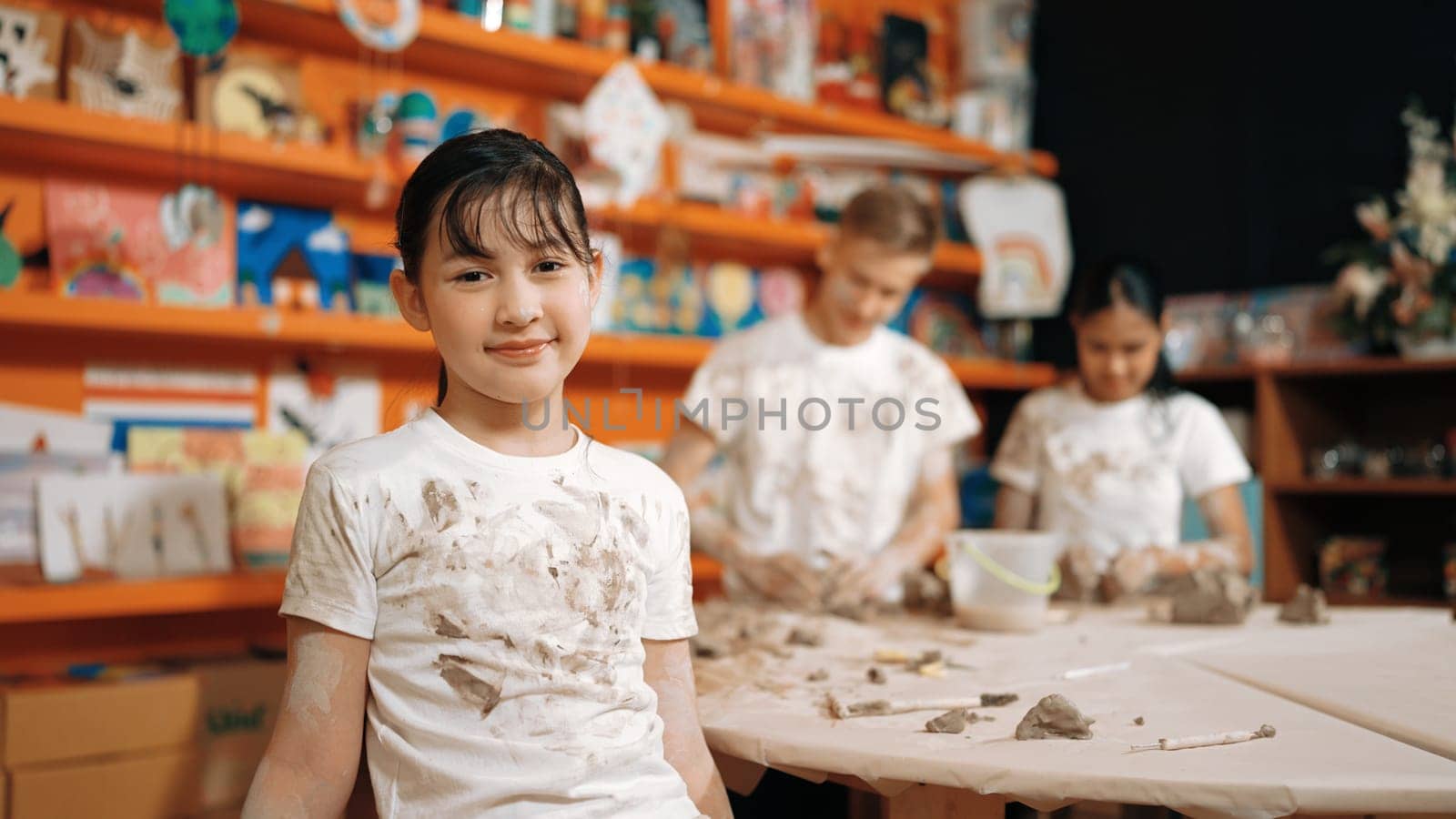 Asian highschool girl look at camera and dirty hand while diverse children modeling clay behind. Happy cute student wearing shirt while looking at camera at workshop. Blurring background. Edification.