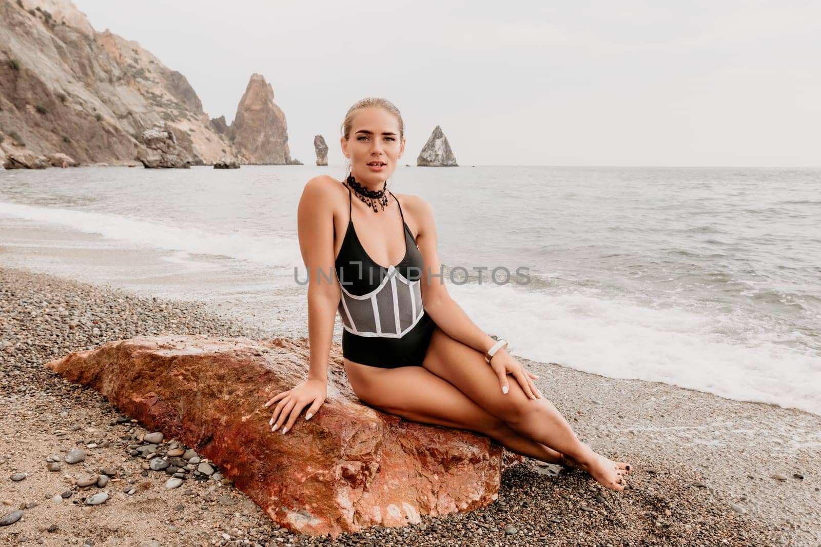 Woman travel portrait. Happy woman with long hair poses on a red volcanic rock at the beach. Close up portrait cute woman in black bikini, smiles at the camera, with the sea in the background. by panophotograph