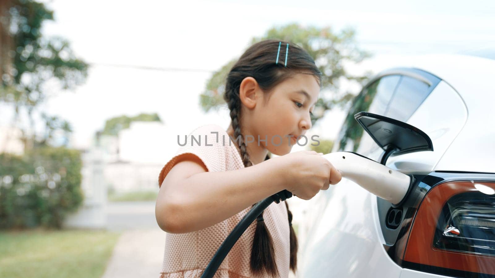 Happy little young girl learn about eco-friendly and energy sustainability as she recharge electric vehicle from home EV charging station. EV car and sustainable future generation concept. Synchronos