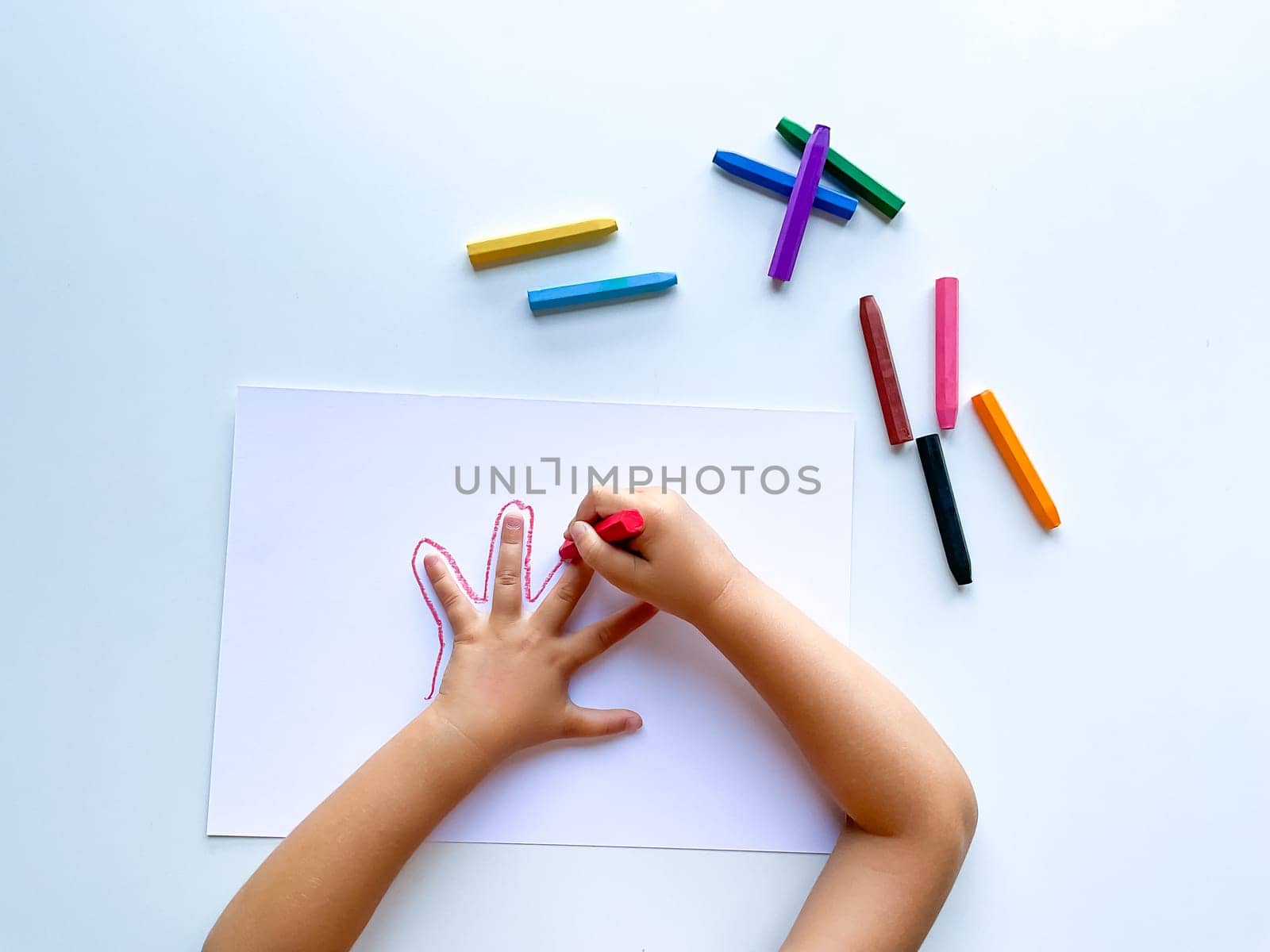 Childrens hands draw their hand with wax crayons on white paper, top view. High quality photo