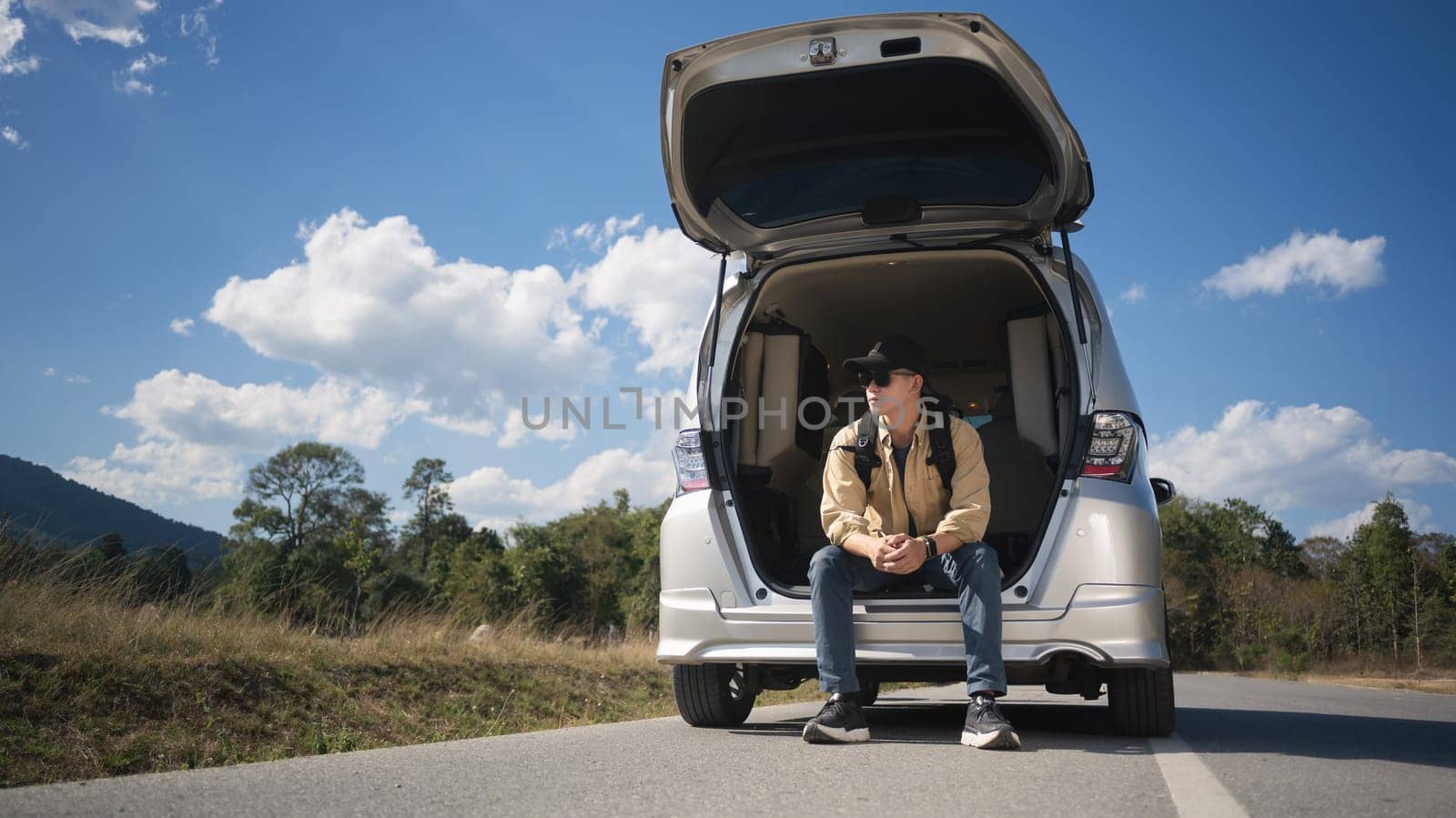 Male traveler on a road trip, sitting on the open trunk of his car against blue sky and white cloud.