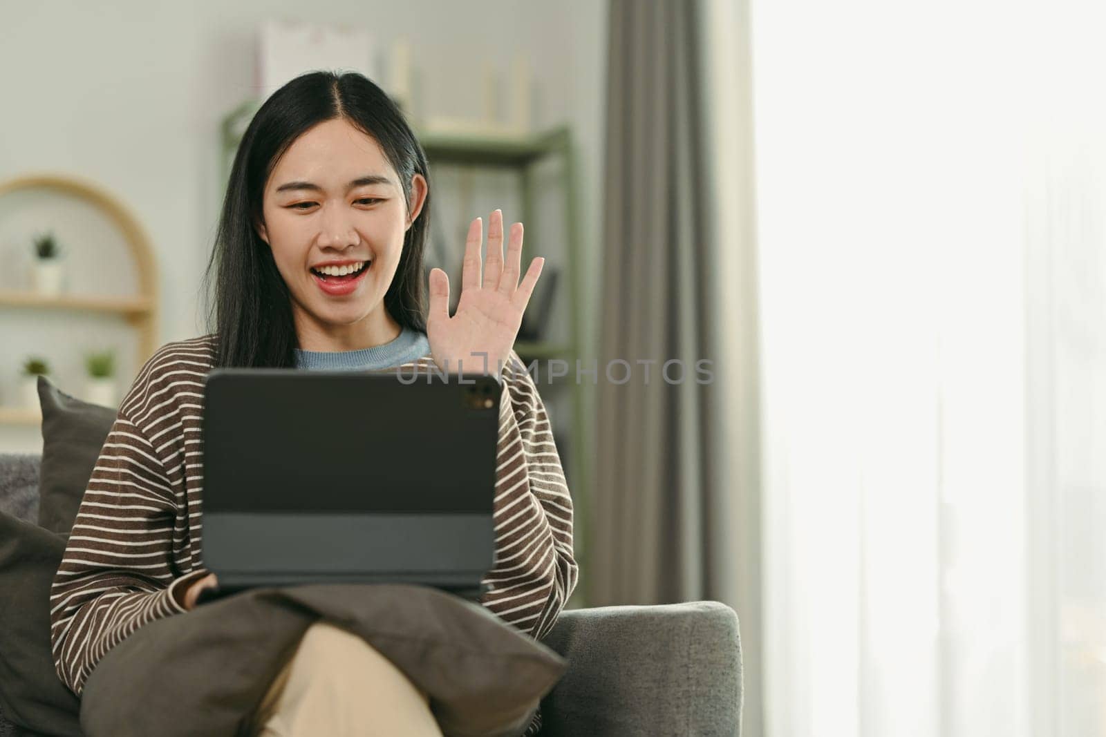 Cheerful young Asian woman smiling and waving hand at digital tablet while chatting on video call.