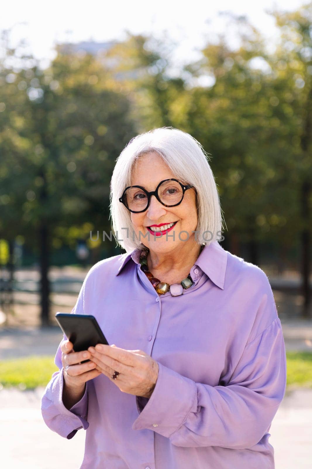 senior woman looking at camera smiling happy using mobile phone outdoors, concept of technology and elderly people leisure, copy space for text