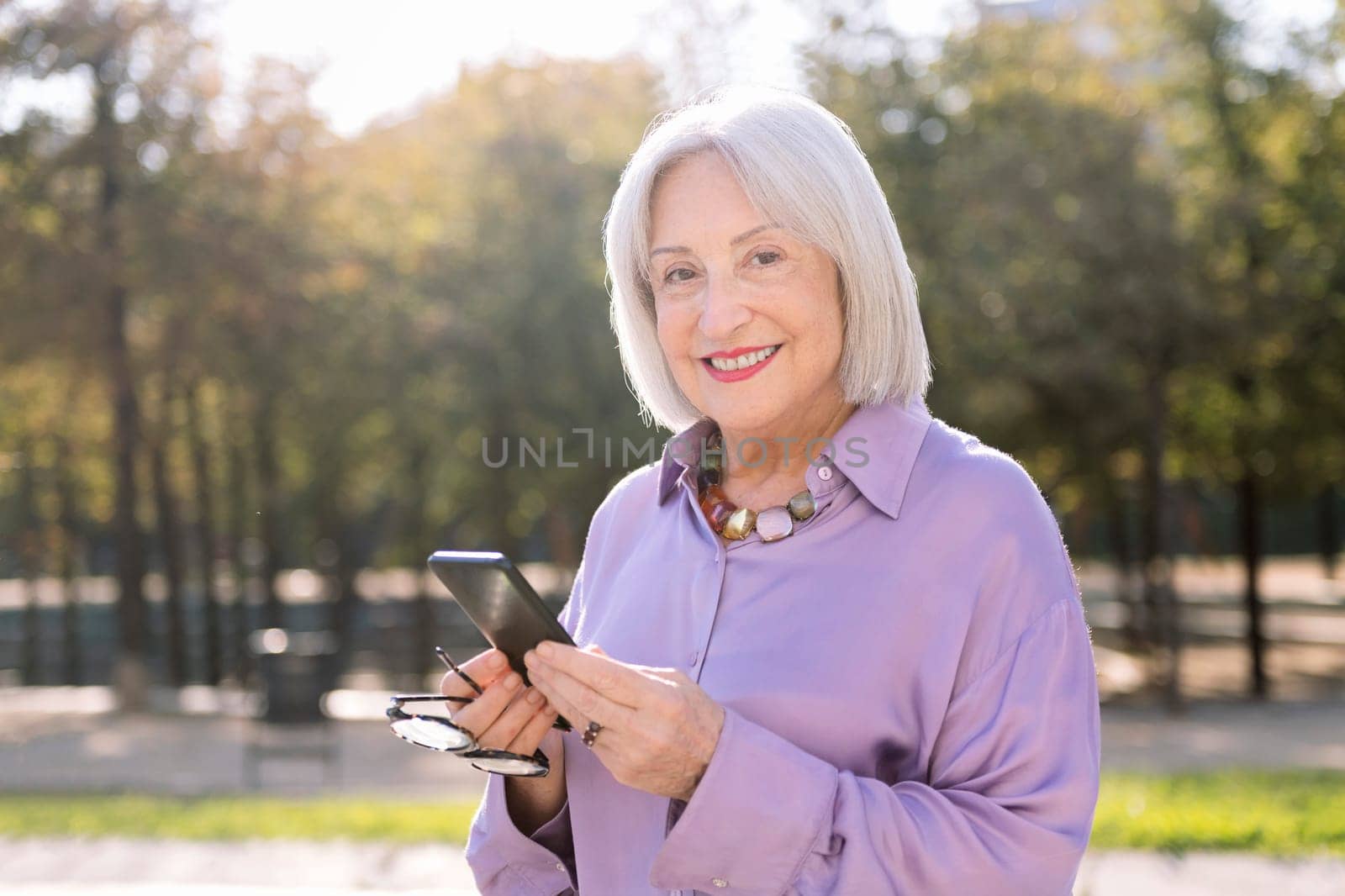 senior woman looking at camera smiling happy using mobile phone outdoors, concept of technology and elderly people leisure, copy space for text