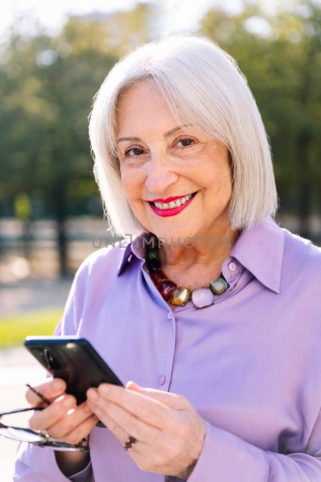 portrait of a senior woman looking at camera smiling happy using mobile phone outdoors, concept of technology and elderly people leisure
