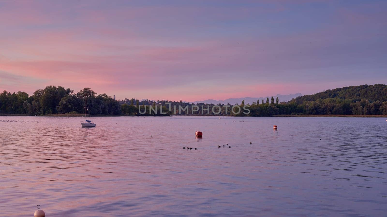 Bodensee Lake Sunrise Panorama. Morning Sunlight Over Tranquil Waters. Witness the mesmerizing dawn over Germany's Bodensee Lake, captured from a boat dock. Embrace the tranquil beauty of the early morning as the sun rises, casting a soft glow on the landscape. The peaceful scene features boats, yachts, and a charming water shack set against a backdrop of a captivating sky. Clouds delicately reflect on the calm water, creating a serene atmosphere. Immerse yourself in the serene beauty of a lakeside sunrise. Explore the harmony of nature, technology, and production as the day unfolds by the lake.