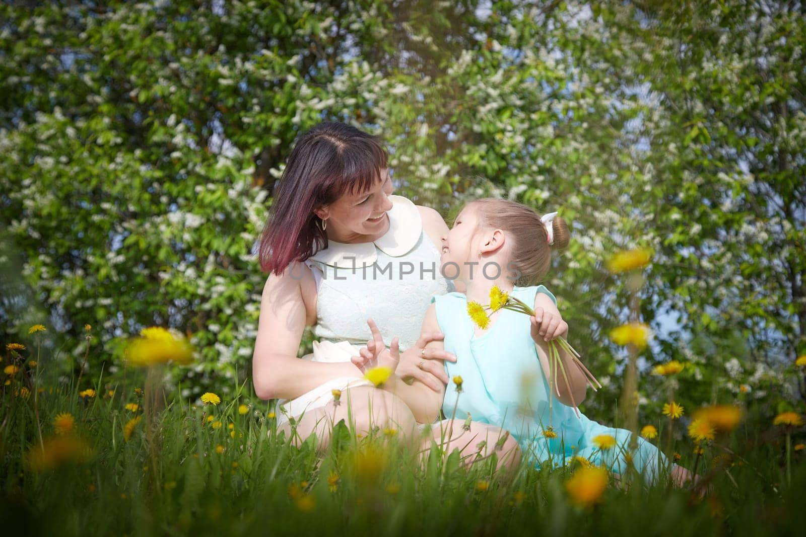 Happy mother and daughter enjoying rest, playing and fun on nature on a green lawn with dandelions and blooming apple tree on background. Woman and girl resting outdoors in summer and spring day