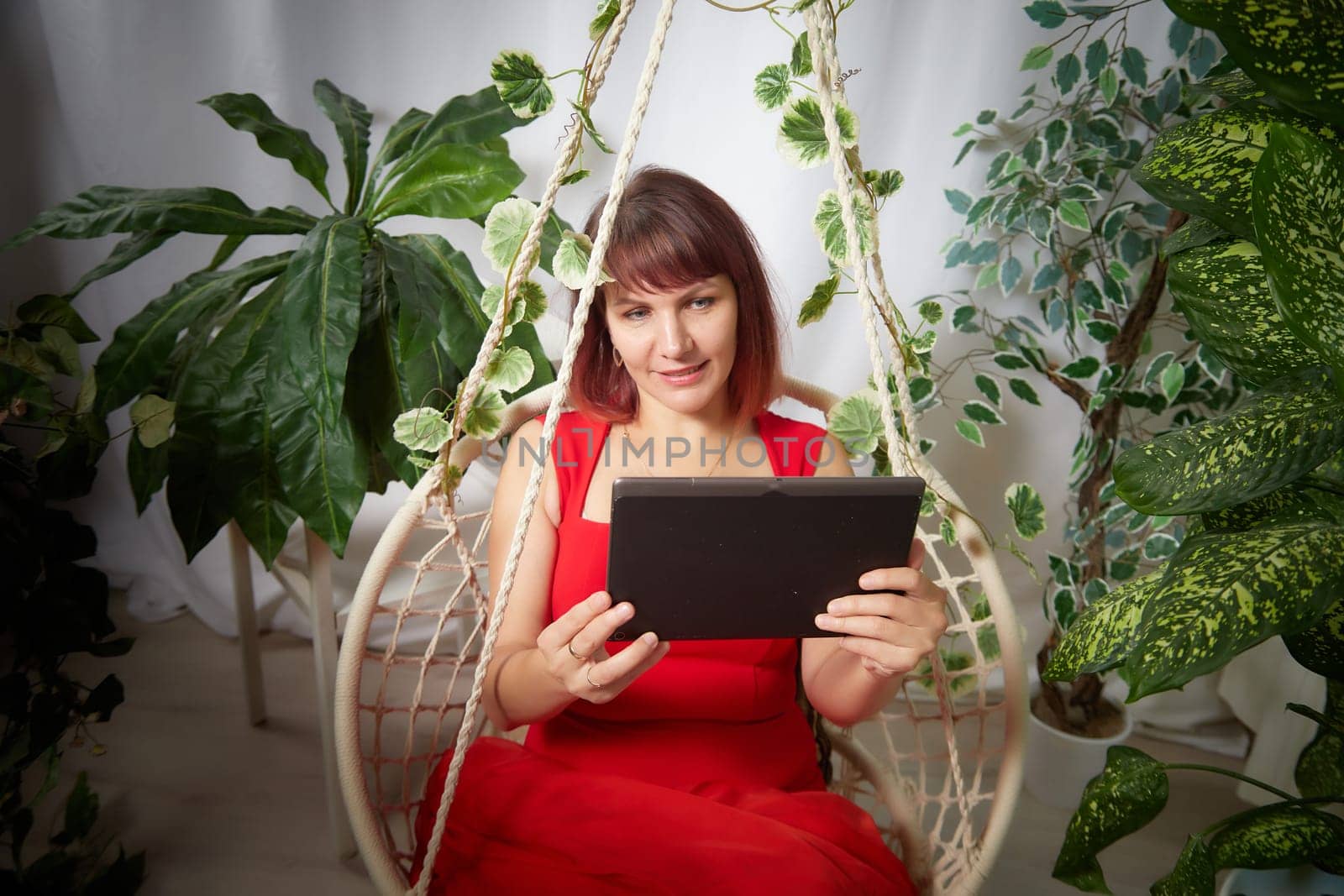 A girl with a laptop and notebook is working on a swing chair in the winter garden. A middle-aged business woman. Psychologist in the office with flowers. Online communication and consultation