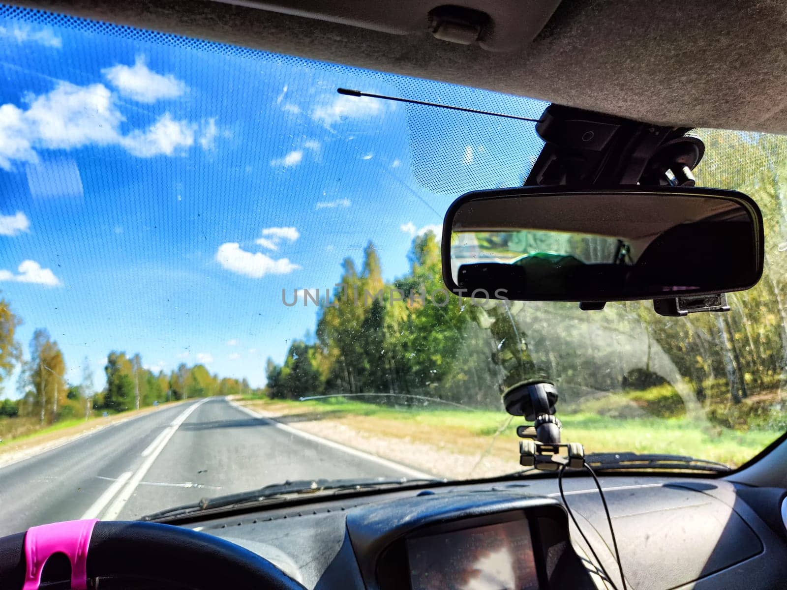 view from a car windshield of natural landscape with road, green trees and blue sky in summer or spring time. Hand of woman on steering wheel. Female traveler driving alone on trip or journey
