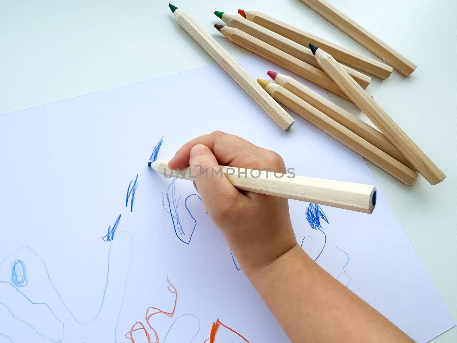 small child draws with colored pencils on paper on white table. by Lunnica