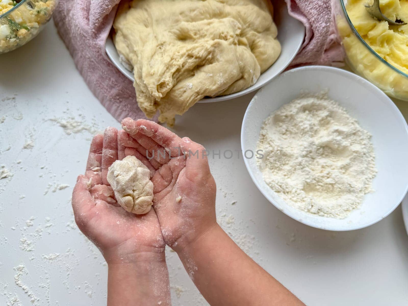 The hands of child knead the dough for making pies on white table, top view. by Lunnica