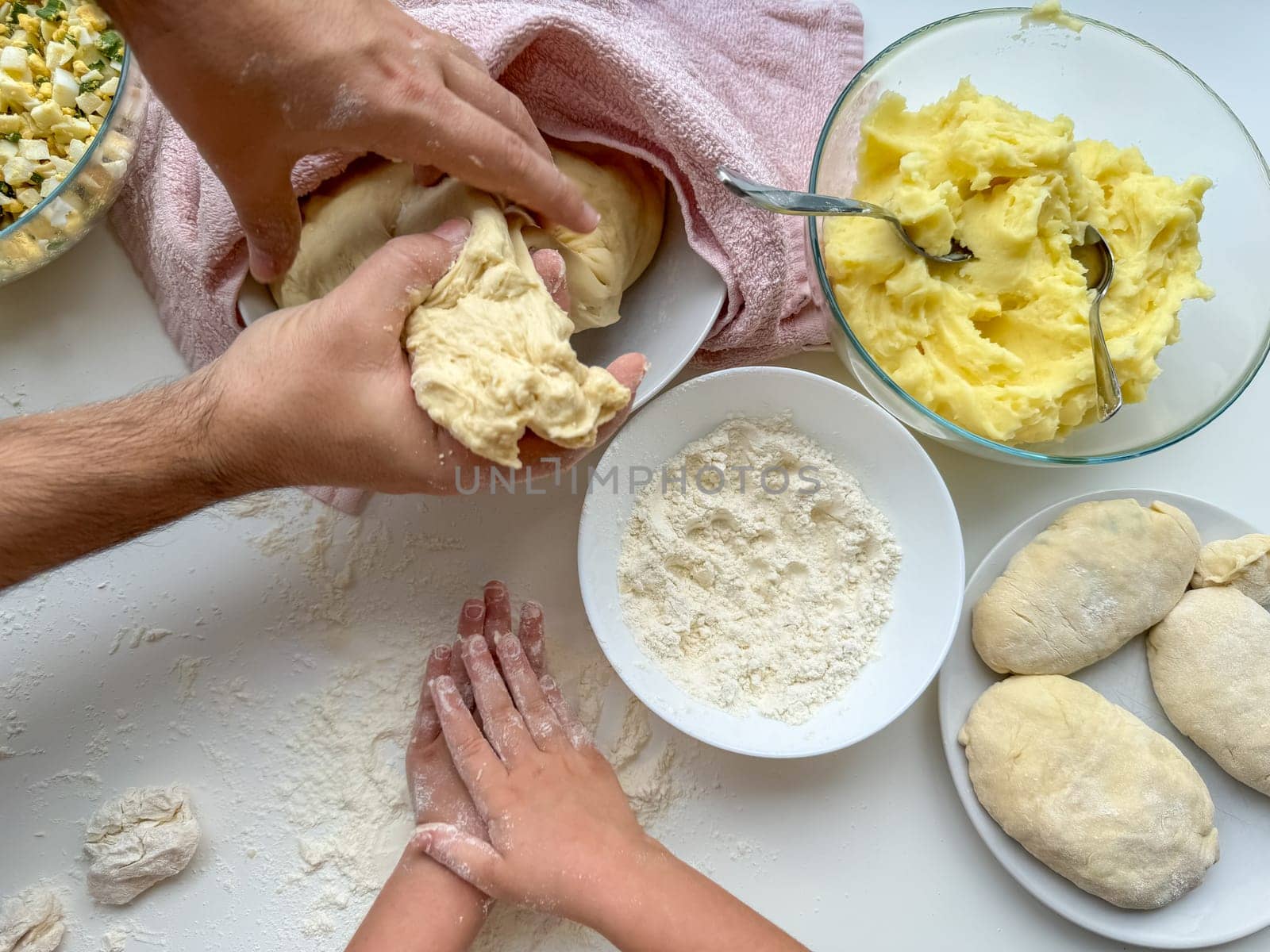 The hands of child and father knead the dough for making pies on white table, top view. High quality