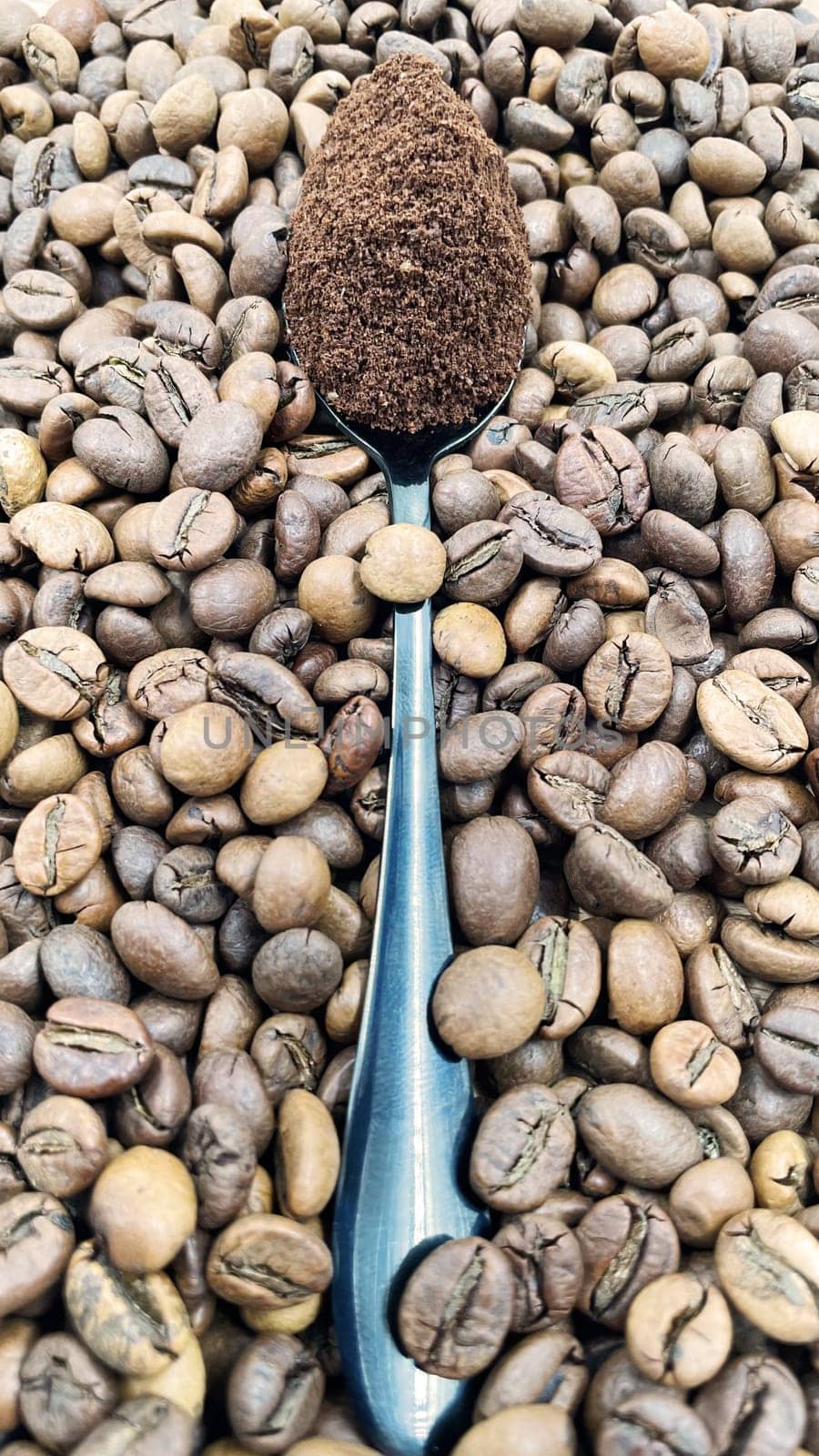 Against the background of roasted aromatic coffee beans lies a metal spoon filled with ground coffee. A drink made from roasted and ground beans from the coffee tree or coffee bush